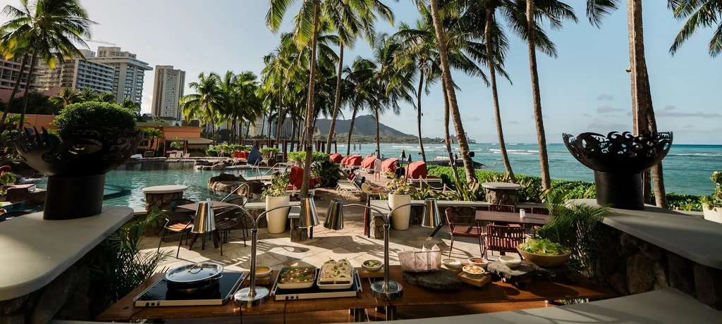Outdoor tropical resort buffet with pool, ocean, palms, and mountain.