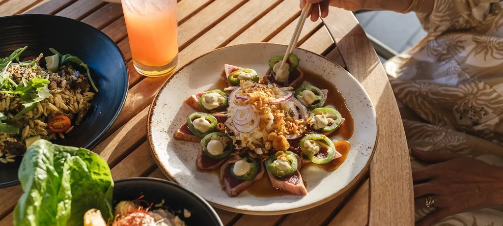 Hand picks seared tuna with chopsticks; salad and drink on wooden table.