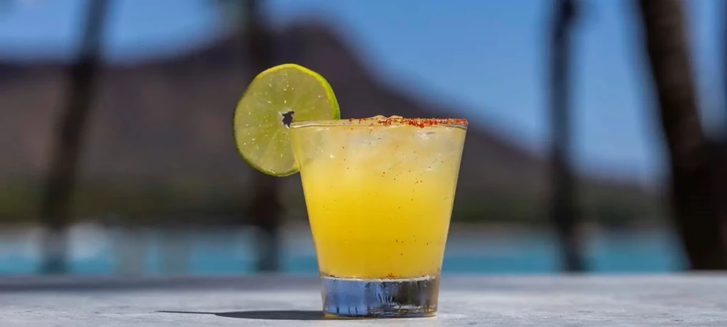 Yellow cocktail with a spiced rim and lime, blurred outdoors.
