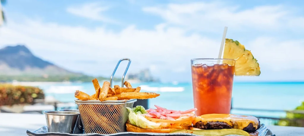 Cheeseburger, fries, tropical drink on a tray, with ocean and mountain.