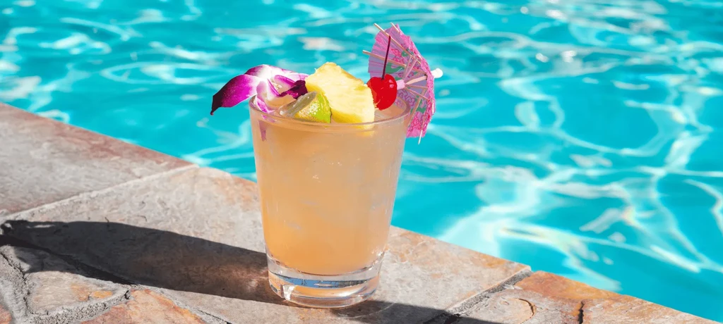 Cocktail with pineapple, purple orchid, umbrella by a clear pool.