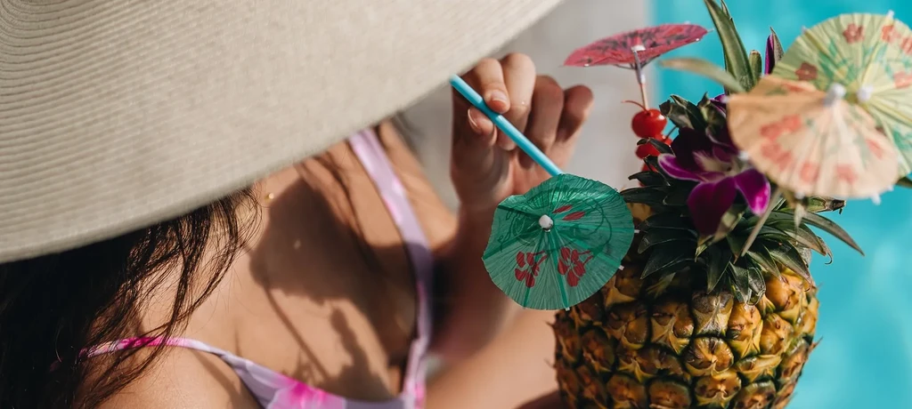 Person sips tropical pineapple cocktail with umbrellas by a pool.