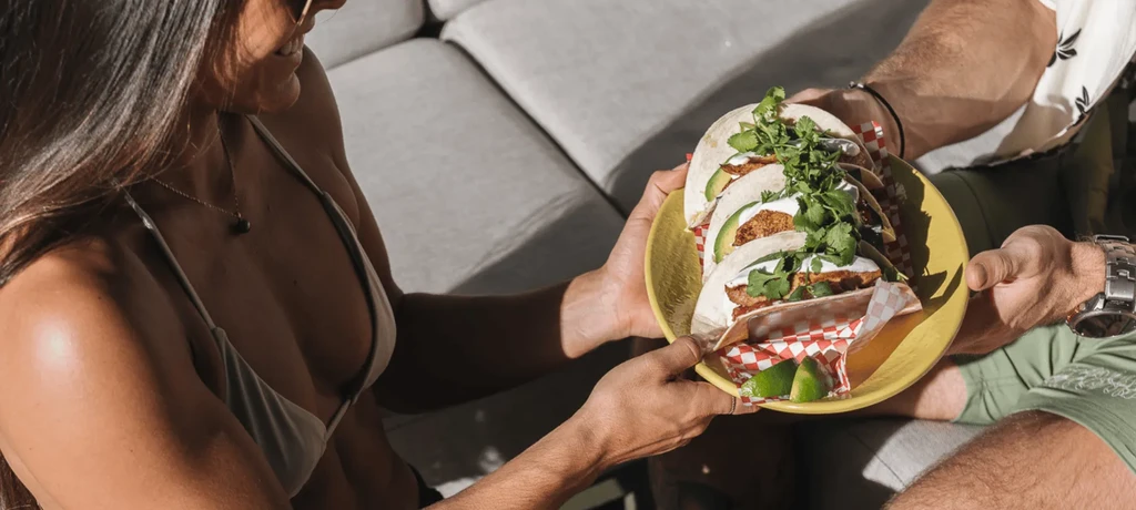 A plate of four loaded tacos passed between two people.