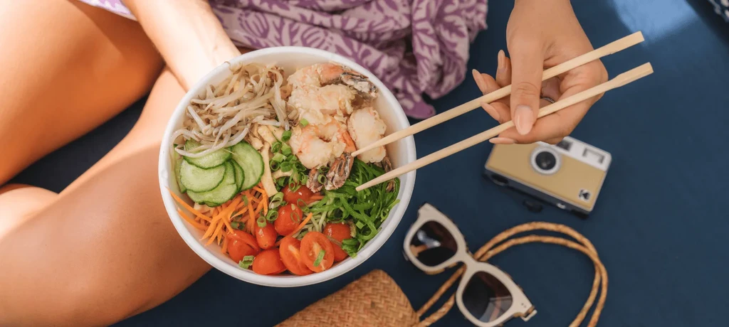 Person holding chopsticks over a colorful poke bowl with shrimp, outdoors.