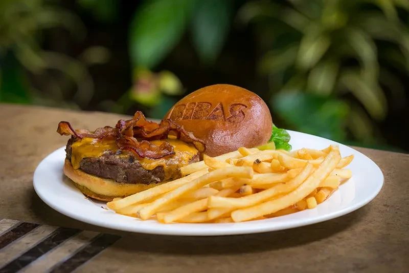 Burger with cheese, bacon, and fries on white plate, blurred green background.