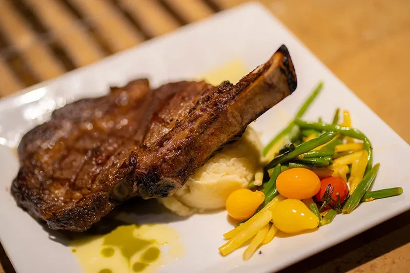 Close-up of grilled bone-in steak, mashed potatoes, colorful vegetables.