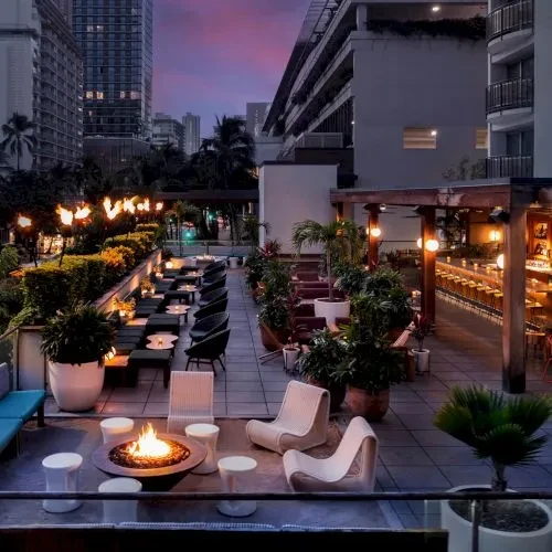 Rooftop bar patio at dusk, showing fire pit, seating, city view.
