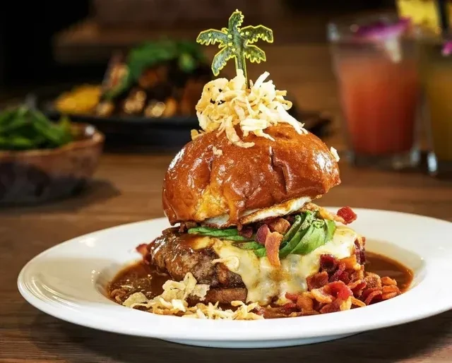 Tall gourmet burger with fried egg, avocado, bacon, sauce, and palm pick.