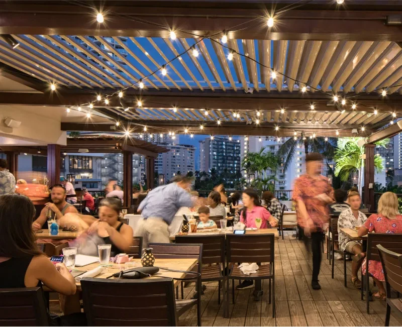 Busy rooftop restaurant at dusk with fairy lights, city views, and diners.