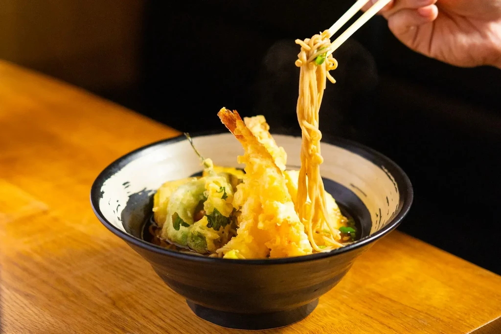Japanese noodles with shrimp tempura, hand lifting noodles from ceramic bowl.
