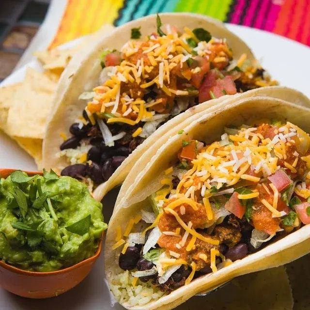 Two soft-shell tacos, guacamole, chips, on a colorful blanket.