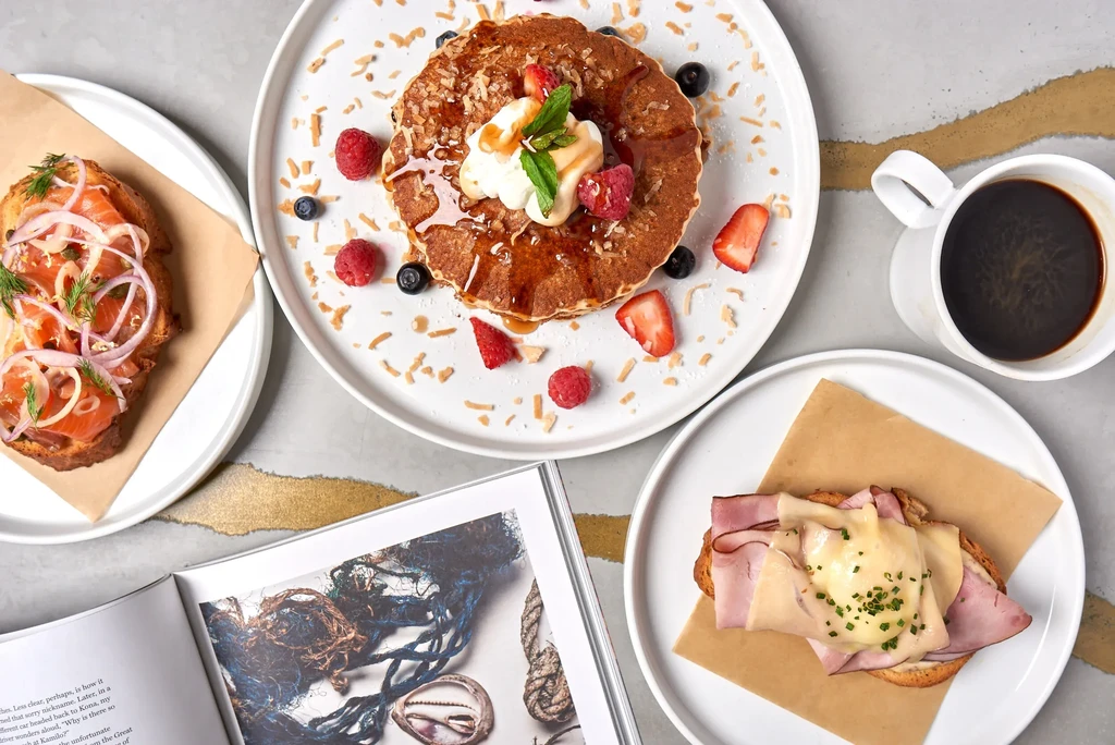 Flat lay: salmon toast, berry pancakes, ham toast, coffee, open book.