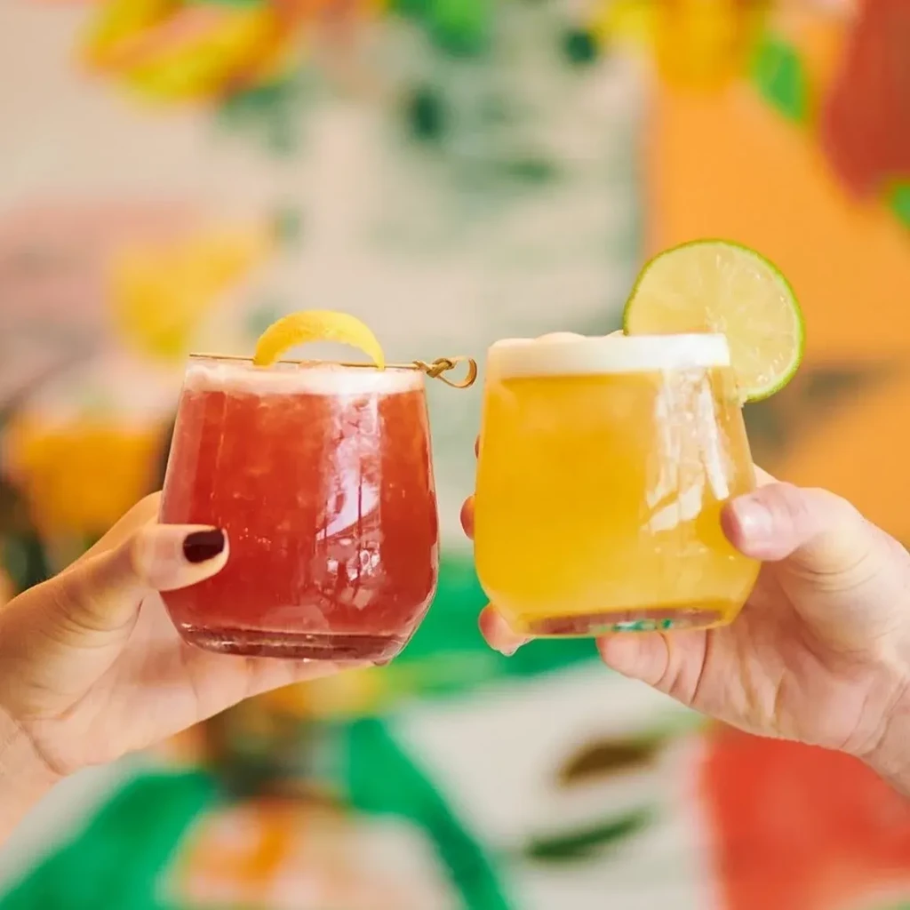 Two hands hold a red and yellow cocktail in a toast.