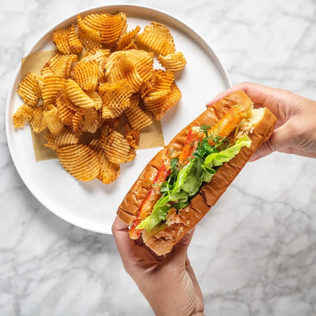 Hands hold a lobster roll with lettuce and waffle chips on marble.