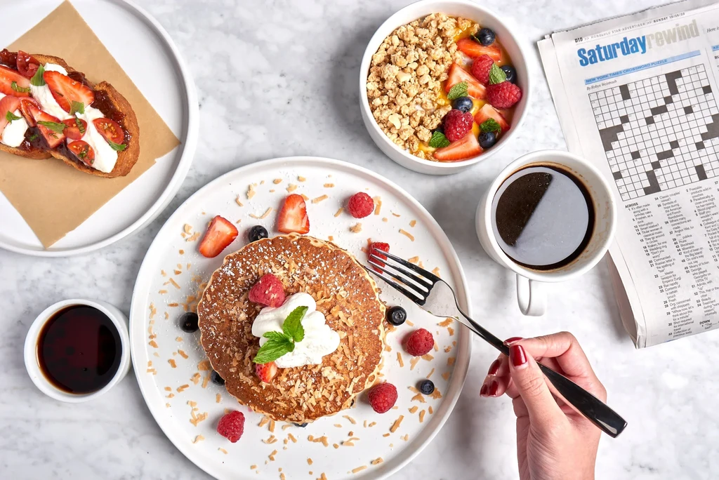 Hand with fork hovers over a colorful breakfast of pancakes, toast, yogurt.