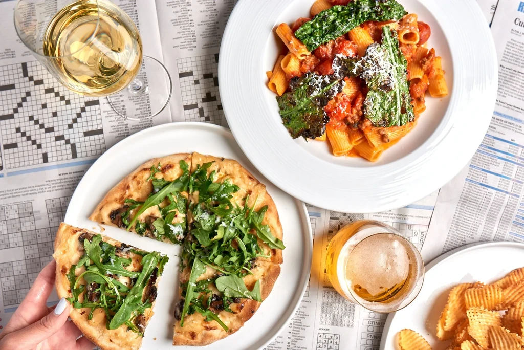 Casual dining spread: pasta, flatbread, chips, wine, and beer on newspaper.