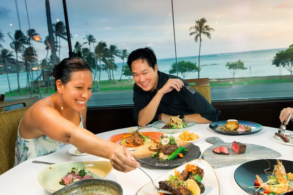 Smiling woman, man in chef's jacket dine with tropical ocean view.