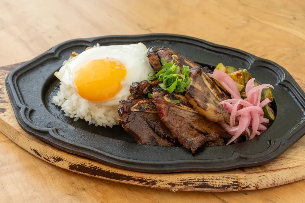 Plated grilled short ribs, rice, sunny egg, pickled onions, green vegetables.