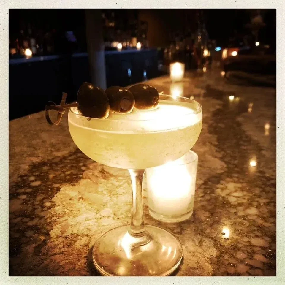A light-colored cocktail with three olives on a skewer at a dimly lit bar.