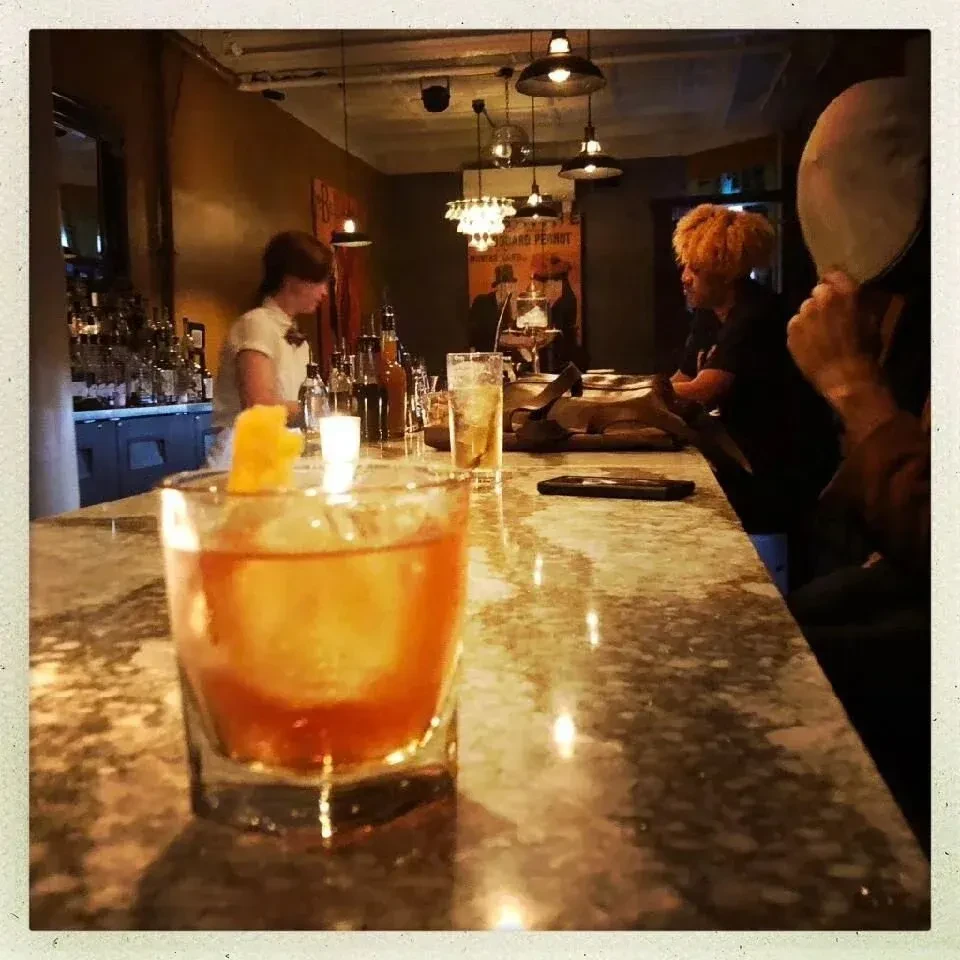 A cocktail sits on a bar counter with a bartender and patrons in background.
