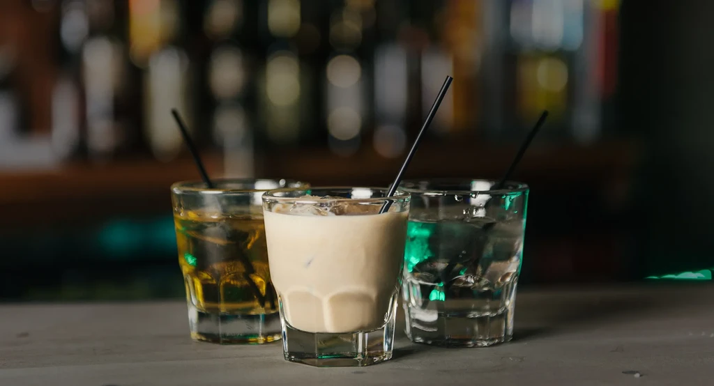 Three amber, beige, clear iced drinks with straws on a bar.