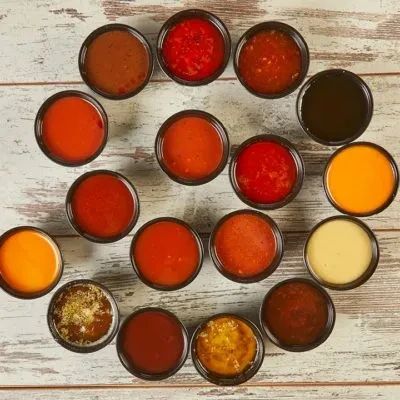 Overhead view of sixteen diverse sauces in dark cups on rustic wood.