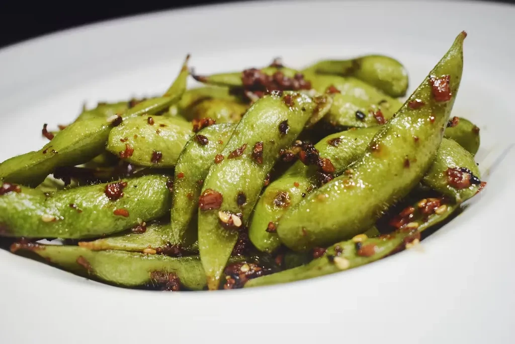 A bowl of green edamame pods with chili flakes and sesame seeds.
