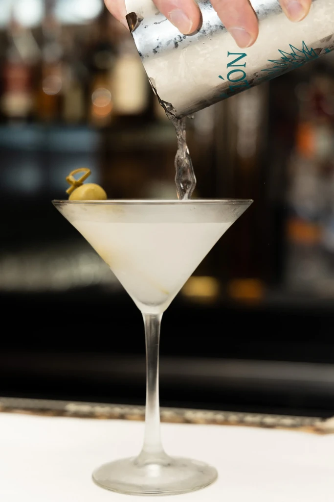 A hand pours a drink from a shaker into a martini glass.