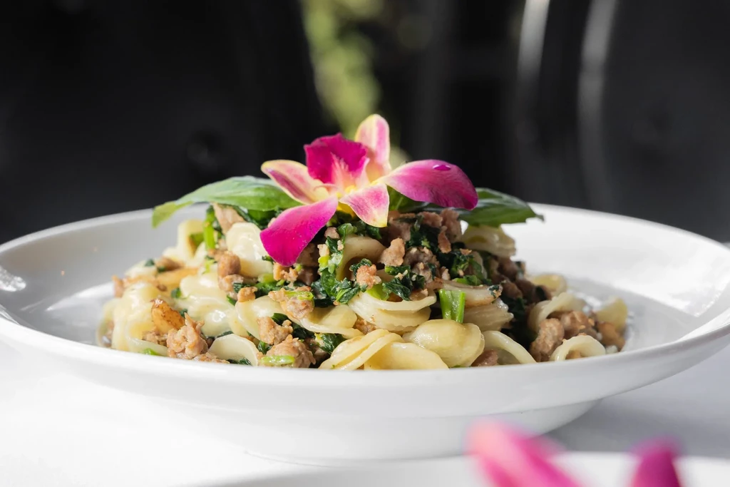 Pasta with meat, greens, and a pink orchid on a white plate.