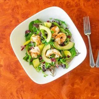 A white square plate of shrimp, avocado, and leafy green salad.