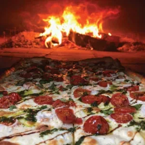A Margherita pizza sits in front of a wood-fired oven with roaring flames.