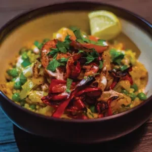 Bowl of shrimp, yellow rice, roasted peppers, peas, garnished with lemon.