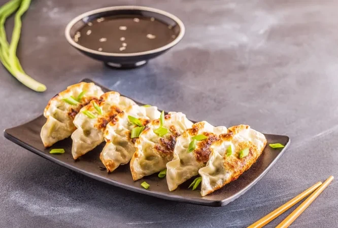 Six crispy gyoza on a black plate with dipping sauce and chopsticks.