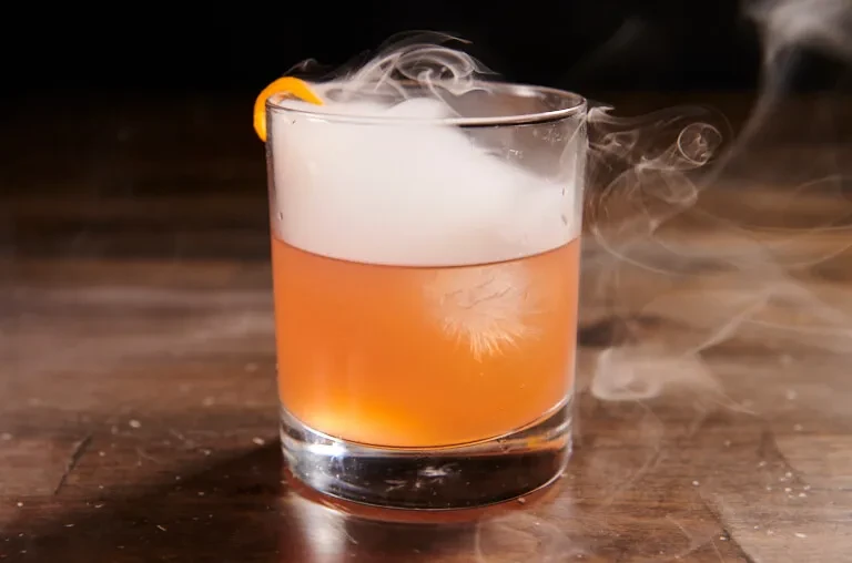 A smoking amber cocktail with an orange peel garnish on a wooden table.