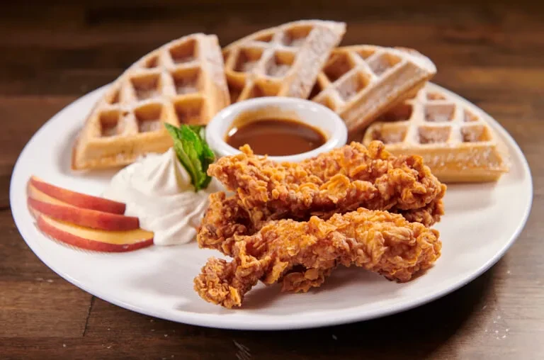 A plate of chicken and waffles with syrup, whipped cream, and peaches.