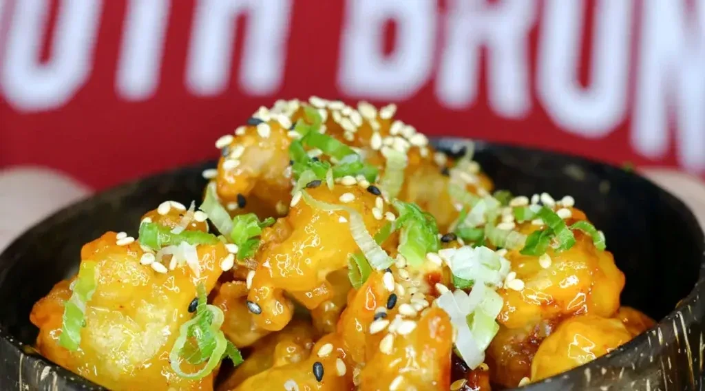Golden glazed cauliflower with sesame seeds and green onions in a dark bowl.