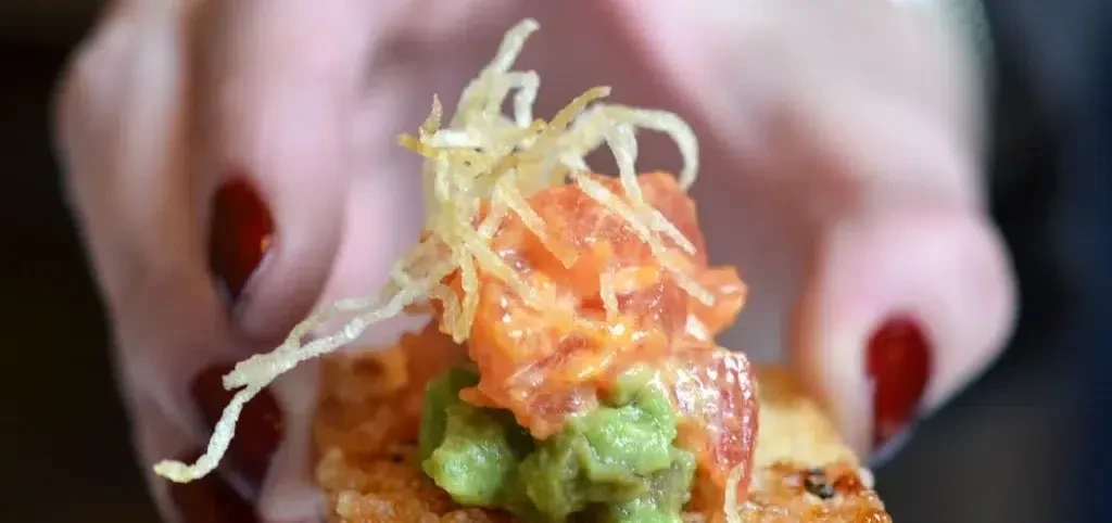 A person holds a small appetizer with salmon, avocado, and crispy toppings.