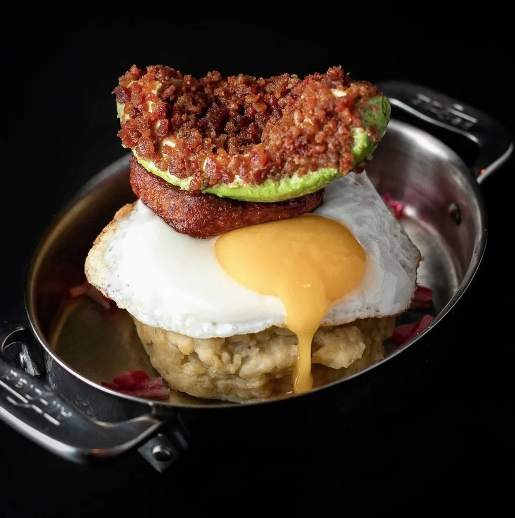A fried egg, sausage, bacon-filled avocado, and grits in a skillet.