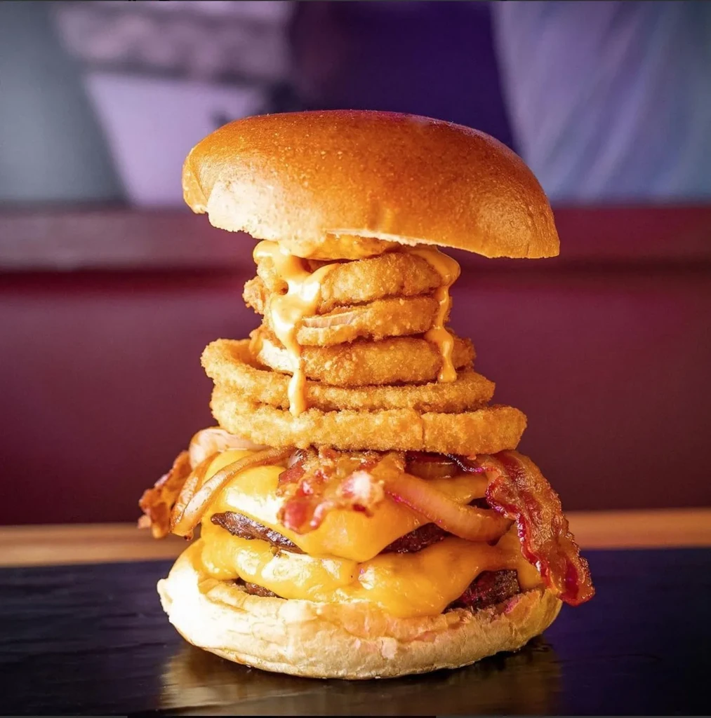 A tall burger with cheese, bacon, grilled onions, and onion rings.