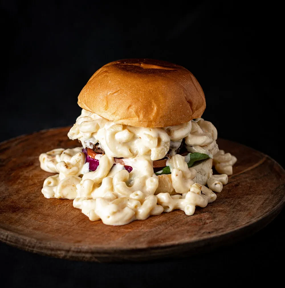 A burger covered in mac and cheese on a wooden board.