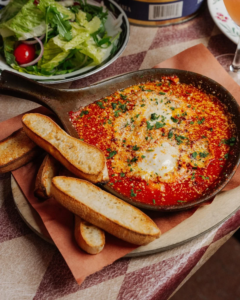 A skillet of baked eggs in tomato sauce with toast and a side salad.