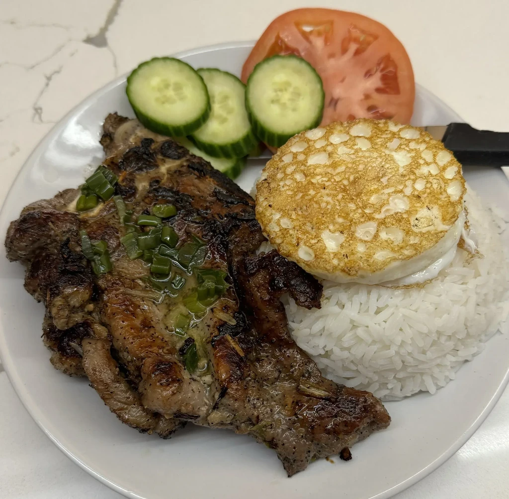 Grilled meat, rice, fried egg, sliced cucumber, and tomato on a plate.