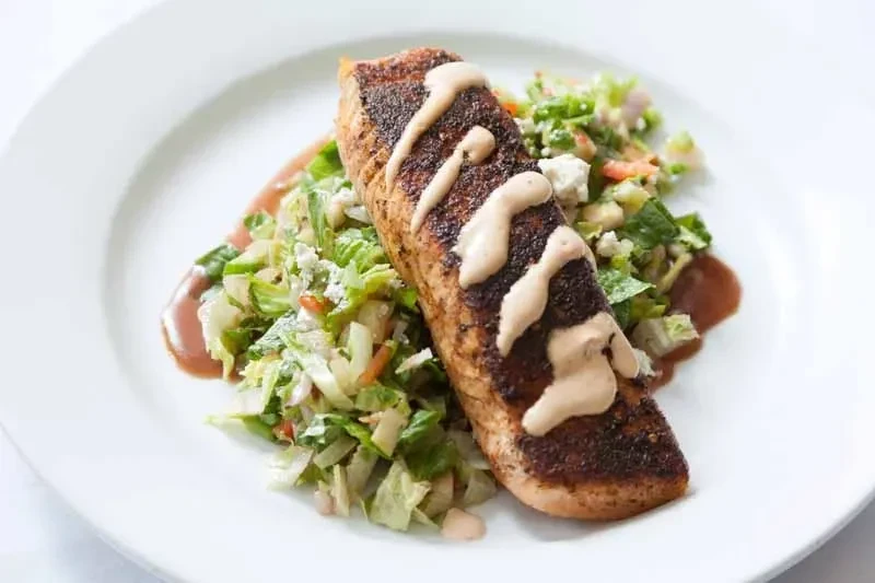 Blackened salmon with drizzled sauce, served with a side salad and dressing.