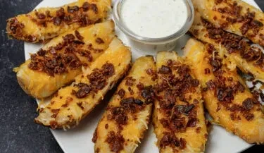 Close-up: Loaded potato wedges with melted cheese, bacon, and white dipping sauce.