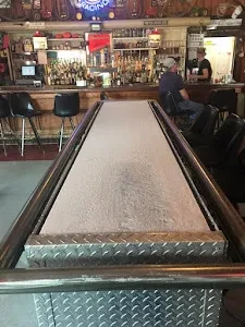 Metallic shuffleboard table in a dimly lit bar with patrons.
