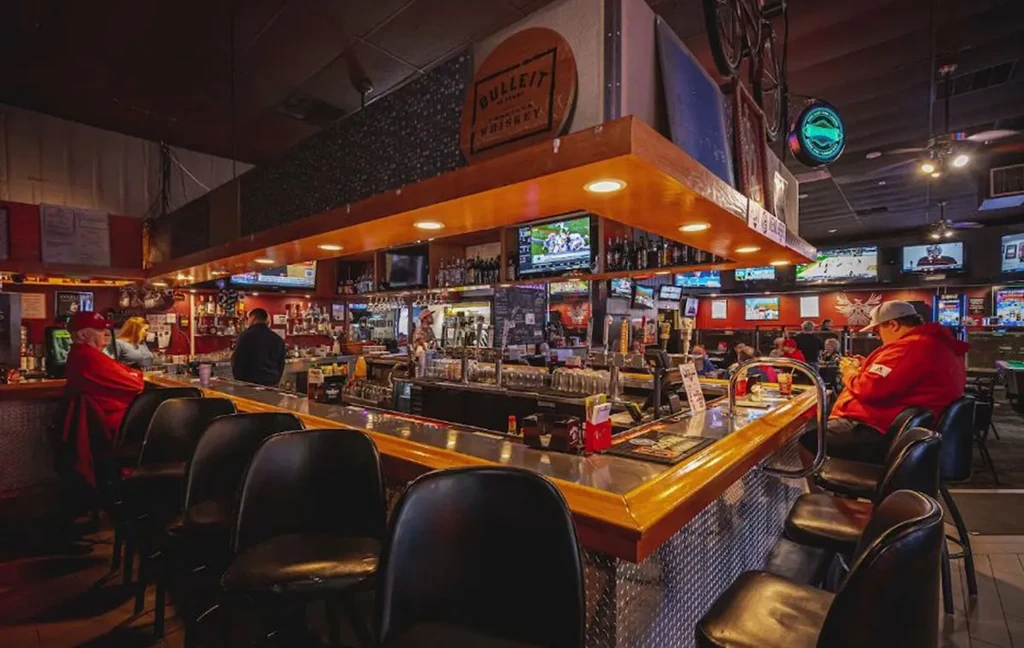 Well-lit sports bar interior with a stocked L-shaped bar and TVs.