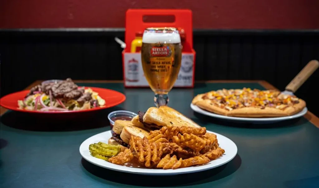 Grilled cheese, waffle fries, salad, pizza, and Stella Artois beer on a table.
