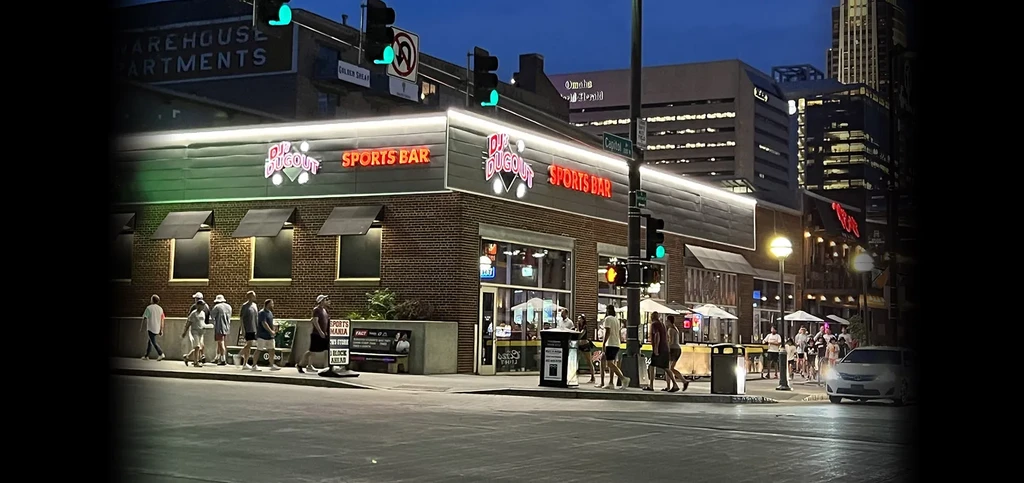 Neon-lit Dugout Sports Bar exterior at night on a lively city corner.