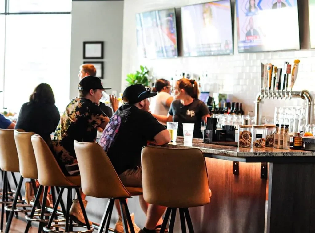 Vibrant modern bar with patrons, a bartender, drinks, and TVs.