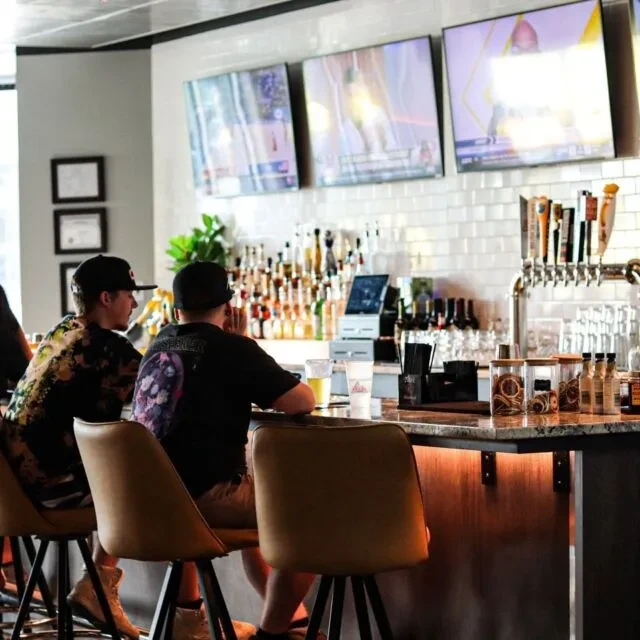 Modern bar interior with a lit counter, bottles, TVs, and two patrons.